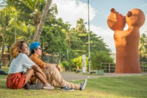 Two travelers relaxing on a lawn beside a cultural sculpture in Brazil